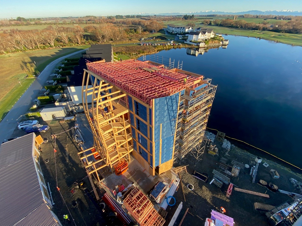 Image of a wooden midrise building under construction next to a small man-made lake.